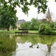 Ruhiger Teich mit Enten im Vordergrund, alte Kirche und gepflegte Gärten im Hintergrund.