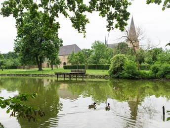 Ruhiger Teich mit Enten im Vordergrund, alte Kirche und gepflegte Gärten im Hintergrund.