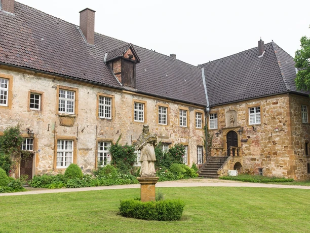 Teil der Klosteranlage Herzebrock mit Blick auf den Eingang des Heimatmuseums Ein historisches Schloss mit verwitterter Fassade und einem gepflegten Garten samt Statue im Vordergrund.