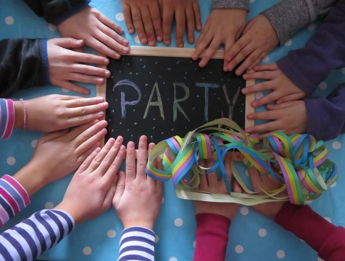 Kinderhände umrahmen eine Tafel mit buntgeschriebenem "PARTY", neben Luftschlangen.