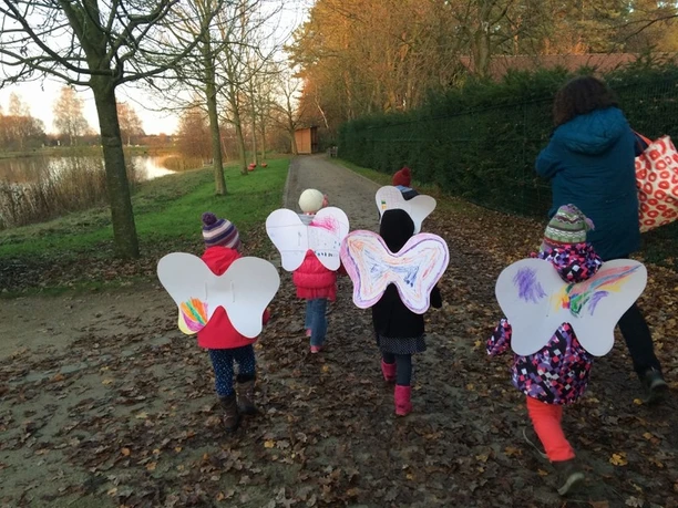 Kinder in bunten Herbstoutfits mit selbstgebastelten Flügeln laufen fröhlich einen Waldweg entlang.