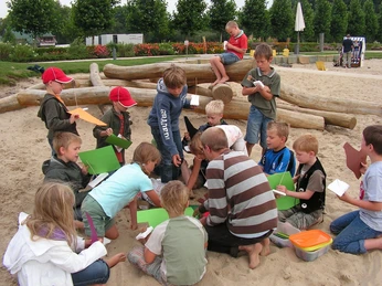 Schiff ahoi! Kinder spielen und basteln auf einem Sandspielplatz mit Holzstämmen und grüner Umrandung.