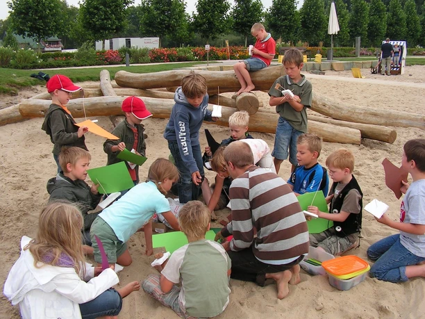 Schiff ahoi! Kinder spielen und basteln auf einem Sandspielplatz mit Holzstämmen und grüner Umrandung.