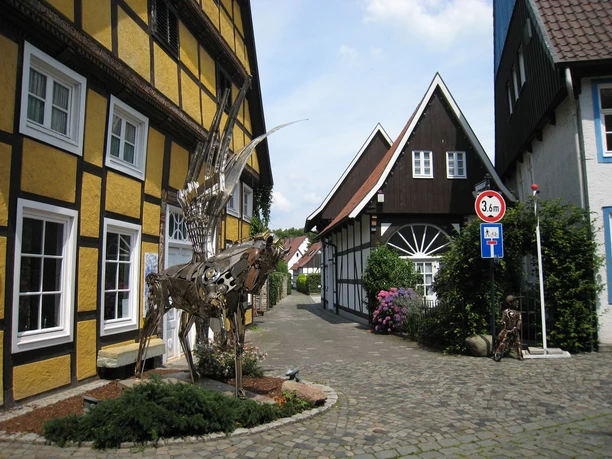 Atelier Monitillo Skulptur eines Hirschs aus Metall in einer malerischen Fachwerkhaus-Straße bei Sonnenschein.