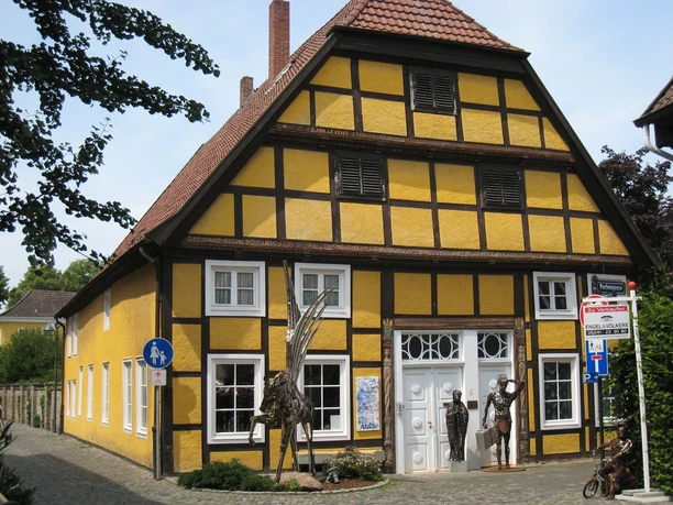 Fachwerkhaus mit gelber Fassade, weißen Fenstern und überdachtem Eingang, umgeben von Bäumen.