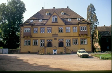 Schloss Amelunxen von 1554 Ockerfarbenes Schloss, zwei Eingänge mit massiven Türen, die mit Spitzrundbogen eingefasst sind, im vorderen Bereich breiter Zuweg und ein parkendes Auto