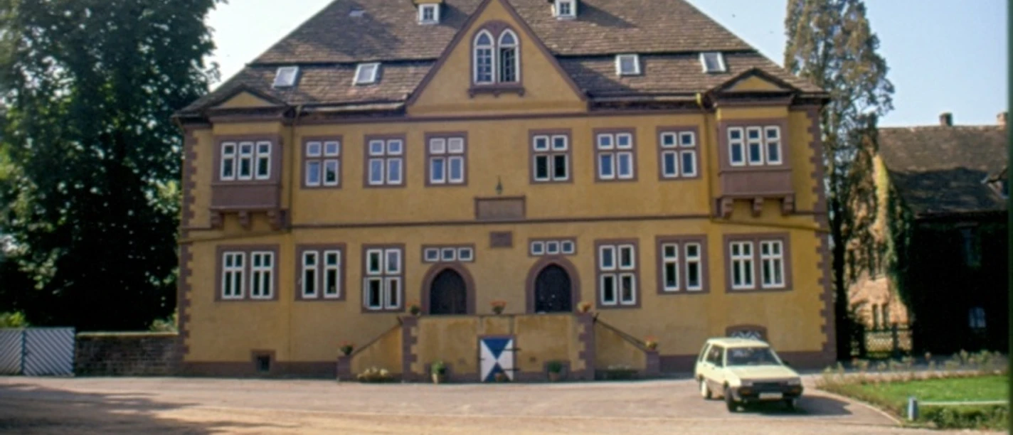 Schloss Amelunxen von 1554 Ockerfarbenes Schloss, zwei Eingänge mit massiven Türen, die mit Spitzrundbogen eingefasst sind, im vorderen Bereich breiter Zuweg und ein parkendes Auto
