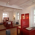 Innenraum einer historischen Ausstellung mit roten Infotafeln und Ausstellungsstücken in Glasvitrinen.
