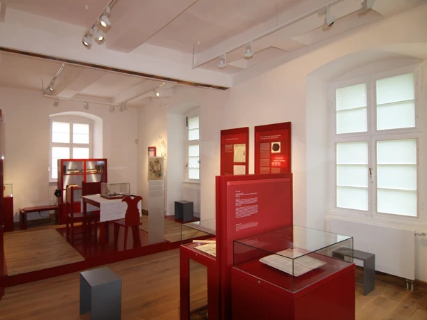Innenraum einer historischen Ausstellung mit roten Infotafeln und Ausstellungsstücken in Glasvitrinen.