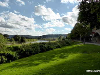 Weserradweg R99 bei Blankenau Grüner Wiesensaum entlang der Weser mit Blick auf bewaldete Hügel, teils bewölkter blauer Himmel.