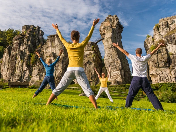 externsteine-horn-bad-meinberg-322-teutoburgerwaldtourismus-dominik-ketz Vier Personen machen Yoga vor den beeindruckenden Felsformationen der Externsteine im Grünen.