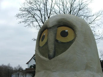 Horst von Hörste - Kopf Skulptur eines stilisierten Eulenkopfes mit großen gelben Augen im Freien vor kahlen Baum.
