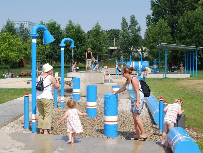 Ein Wasserspielplatz mit blauen Röhren, Kindern und Erwachsenen beim Spielen und Erkunden im Freien.