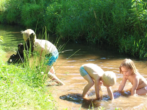 Wasserspielplatz Parkteil Nord Drei Kinder spielen fröhlich im seichten Wasser eines kleinen, mit Gras umgebenen Bachs.