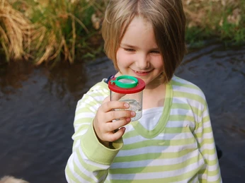Wasserforscher Ein Kind im gestreiften Pullover hält lächelnd ein Becherglas mit einem Fang, neben einem Bach.