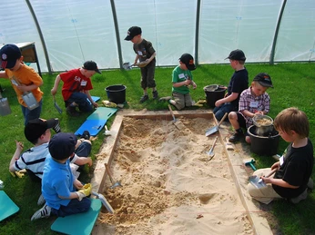 Archäologiefeld Kinder spielen mit Schaufeln im Sandkasten eines Gewächshauses, fröhliche, gemeinschaftliche Umgebung.