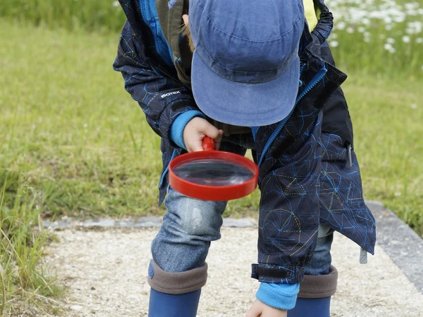 Detektivarbeit Kind in blauer Jacke und Gummistiefeln untersucht mit einer Lupe den Boden auf einem Kiesweg.