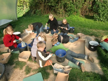 Arbeiten wie Archäologen Kinder spielen im Freien auf einer Sandfläche, umgeben von Grün; sie sind in Aktivitäten vertieft.