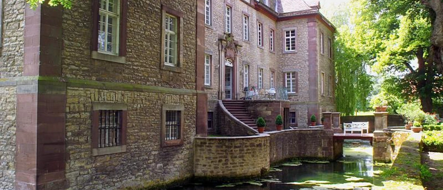 Schloss Welda Altes Schloss mit Sandsteinfassade, umgeben von Wassergraben und Bäumen, reflektierend im Wasser.