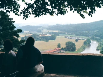 Zwei Personen sitzen auf einer Bank mit Blick auf eine weite, bewaldete Landschaft und einen Fluss.