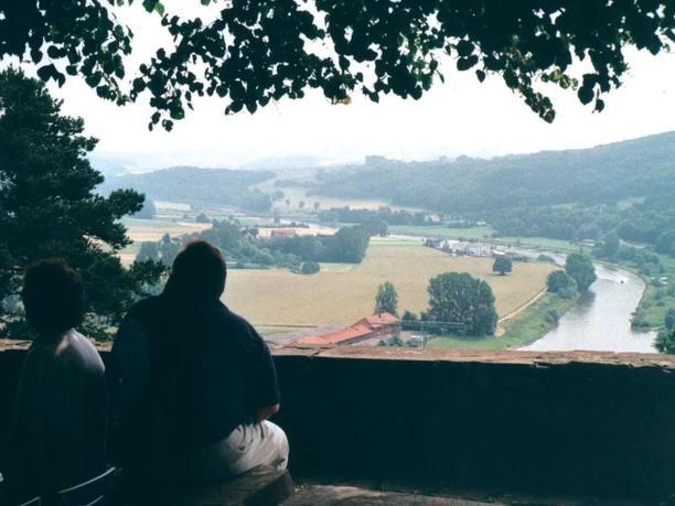 Burg Aussicht Zwei Personen sitzen auf einer Bank mit Blick auf eine weite, bewaldete Landschaft und einen Fluss.