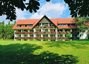 Außenansicht HolsingVital Dreigeschossiges Gebäude mit roten Dachziegeln, Balkonen voller roter Blumen, umgeben von Grün.
