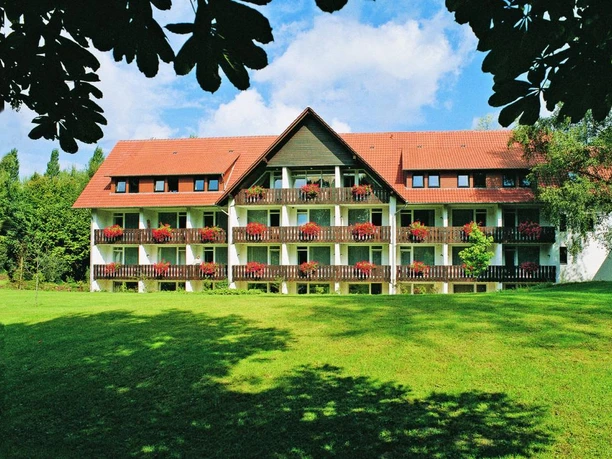Außenansicht HolsingVital Dreigeschossiges Gebäude mit roten Dachziegeln, Balkonen voller roter Blumen, umgeben von Grün.