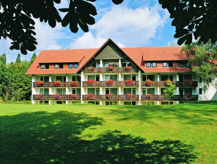 Außenansicht HolsingVital Dreigeschossiges Gebäude mit roten Dachziegeln, Balkonen voller roter Blumen, umgeben von Grün.