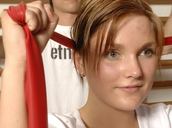 Übung Theraband HolsingVital Zwei junge Personen üben mit farbigen Therabändern vor einer Sprossenwand in einem Fitnessraum.