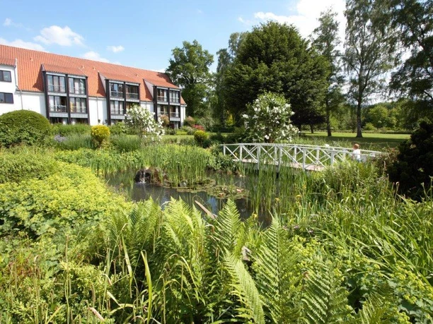 Landschaftspark HolsingVital Ein üppiger Garten mit Teich und Holzbrücke, umgeben von einem Gebäude mit rotem Ziegeldach.