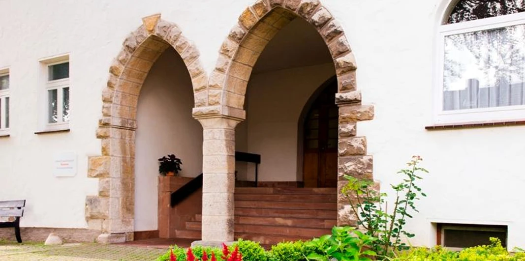 Zwei steinerne Rundbogenportale in einer weißen Fassade, umgeben von gepflegten Blumenbeeten.