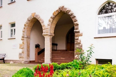 Zukunftswerkstatt Ökumene Warburg-Germete Zwei steinerne Rundbogenportale in einer weißen Fassade, umgeben von gepflegten Blumenbeeten.