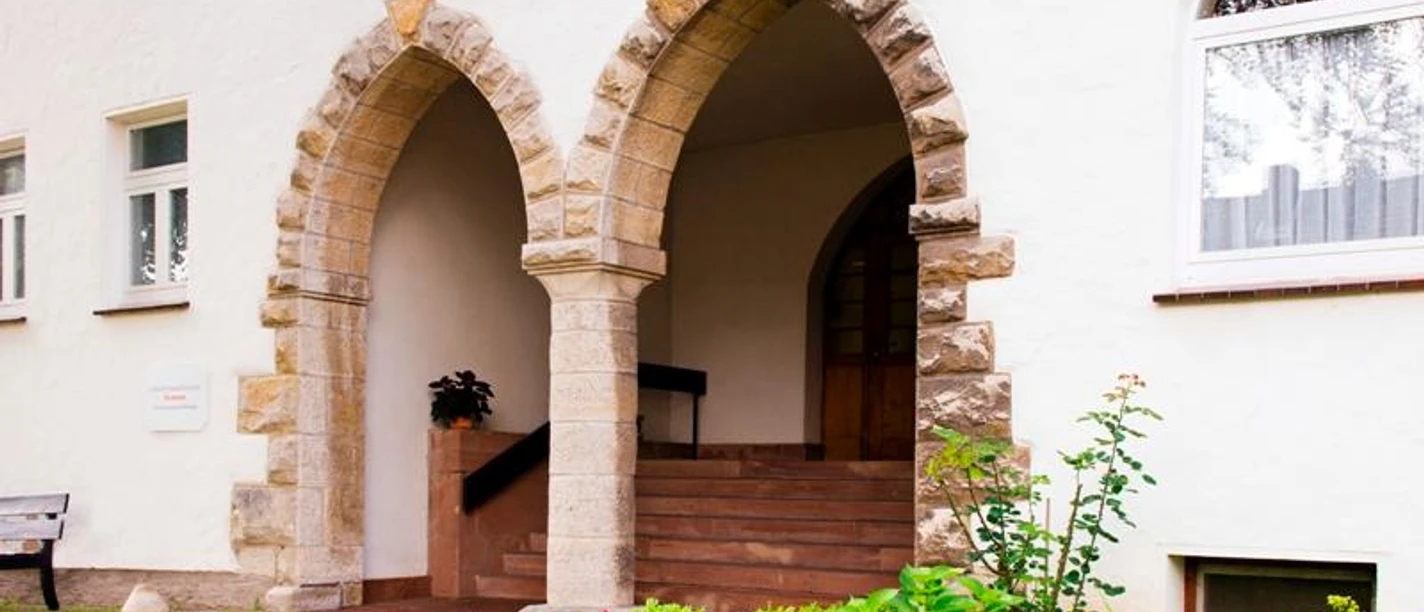 Zukunftswerkstatt Ökumene Warburg-Germete Zwei steinerne Rundbogenportale in einer weißen Fassade, umgeben von gepflegten Blumenbeeten.