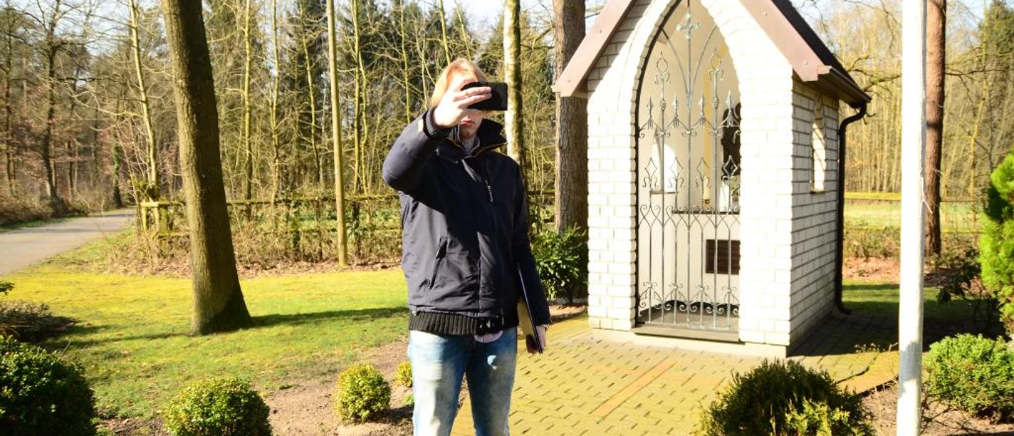 Heiligenhäuschen am Wapelweg Ein Mann macht ein Selfie vor einem kleinen, weißen Heiligenhäuschen im sonnigen Wald.