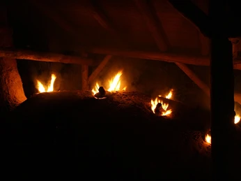 Holzkohlemeiler bei Nacht mit lodernden Flammen und ruhiger Holzkonstruktion im Hintergrund.