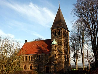 Historische Kirche Hagedorn aus Stein mit rotem Dach, umgeben von Bäumen unter blauem Himmel.