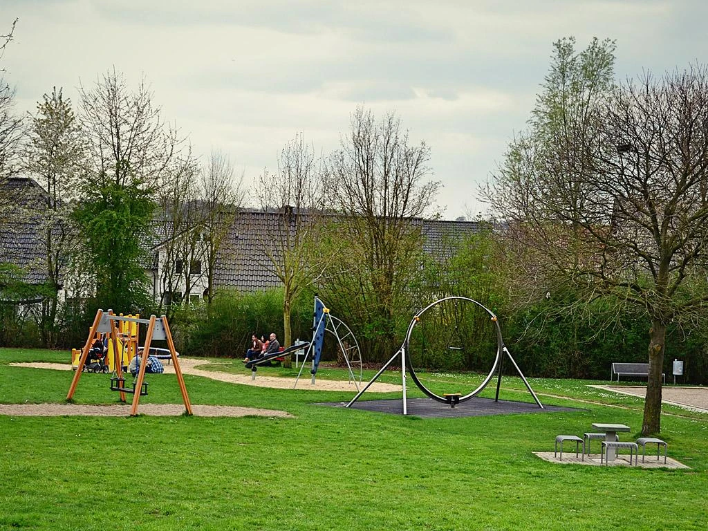 Spielplatz mit Schaukeln und Rutschen im Mehrgenerationenpark, umgeben von grüner Wiese und Bäumen.