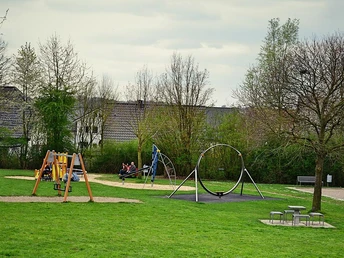 Spielplatz mit Schaukeln und Rutschen im Mehrgenerationenpark, umgeben von grüner Wiese und Bäumen.
