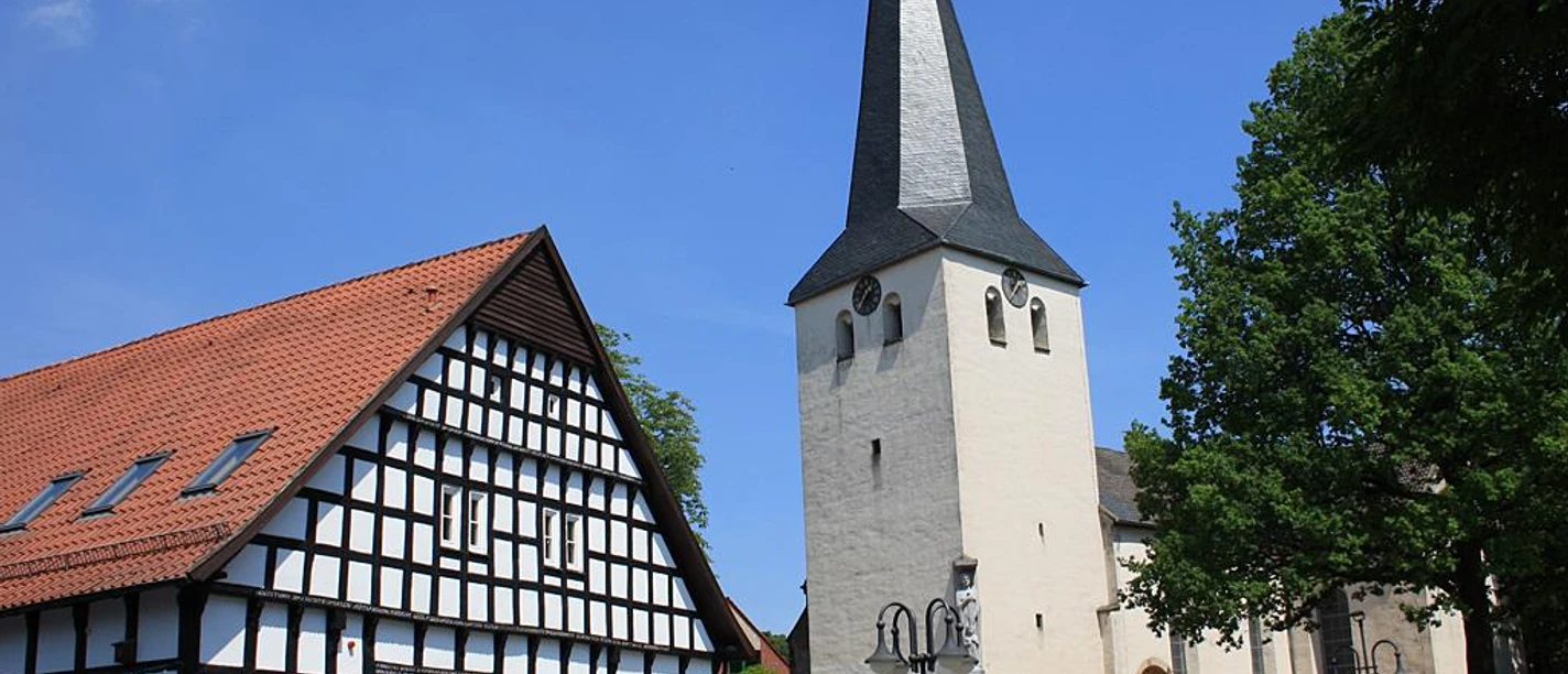 Historische Laurentiuskirche mit markantem spitzen Turm neben einem Fachwerkhaus, blauer Himmel.