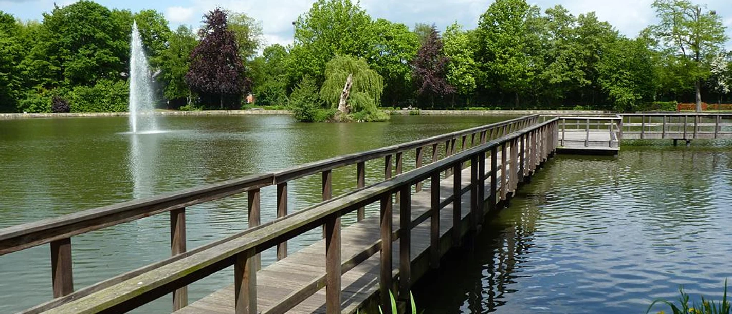Holzsteg überquert einen Teich mit Fontäne im Steinmeisterpark, umgeben von Bäumen und blauem Himmel.