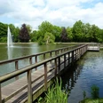Steinmeisterpark Holzsteg überquert einen Teich mit Fontäne im Steinmeisterpark, umgeben von Bäumen und blauem Himmel.