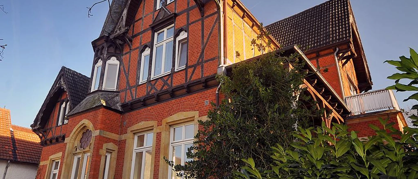 Rote Backsteinvilla mit Fachwerkgiebel, weißen Fenstern und umgeben von grünen Büschen und Bäumen.