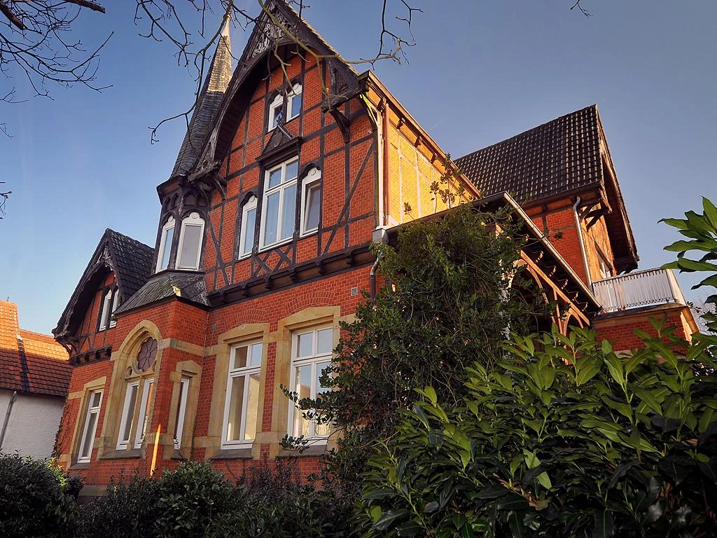 Villa Grüter Rote Backsteinvilla mit Fachwerkgiebel, weißen Fenstern und umgeben von grünen Büschen und Bäumen.