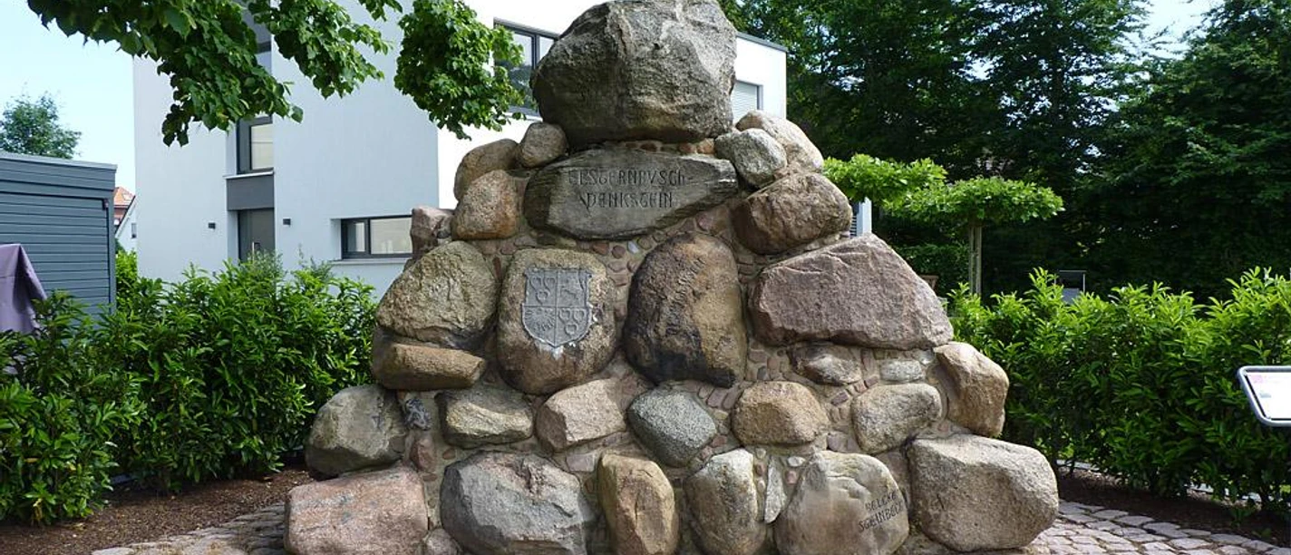 Steinhaufen-Denkmal aus glatten, großen Felsen, umgeben von grünen Hecken und Bäumen.