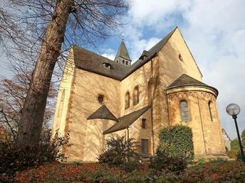 Stiftskirche mit runden Türmen und Spitzgiebel, umgeben von Bäumen und Pflanzen, bei strahlendem Himmel.