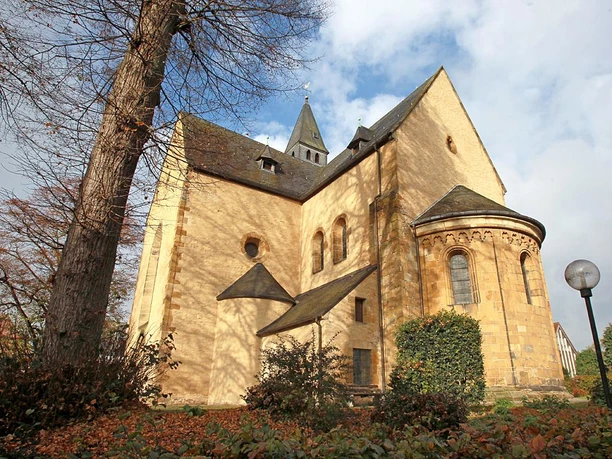Stiftskirche mit runden Türmen und Spitzgiebel, umgeben von Bäumen und Pflanzen, bei strahlendem Himmel.