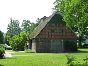 Ein historisches Backsteingebäude mit großen Holztoren, umgeben von grünen Bäumen und Wiesen.