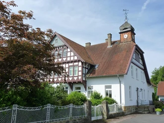 Fachwerkhaus mit roten Ziegeldach, Turmuhr, umgeben von grünen Büschen und einem malerischen Zaun.