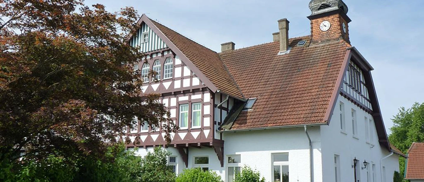 Fachwerkhaus mit roten Ziegeldach, Turmuhr, umgeben von grünen Büschen und einem malerischen Zaun.