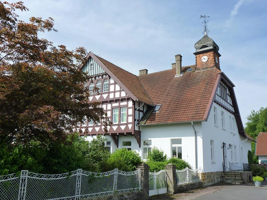 Sattelmeierhof Ringsthof Fachwerkhaus mit roten Ziegeldach, Turmuhr, umgeben von grünen Büschen und einem malerischen Zaun.